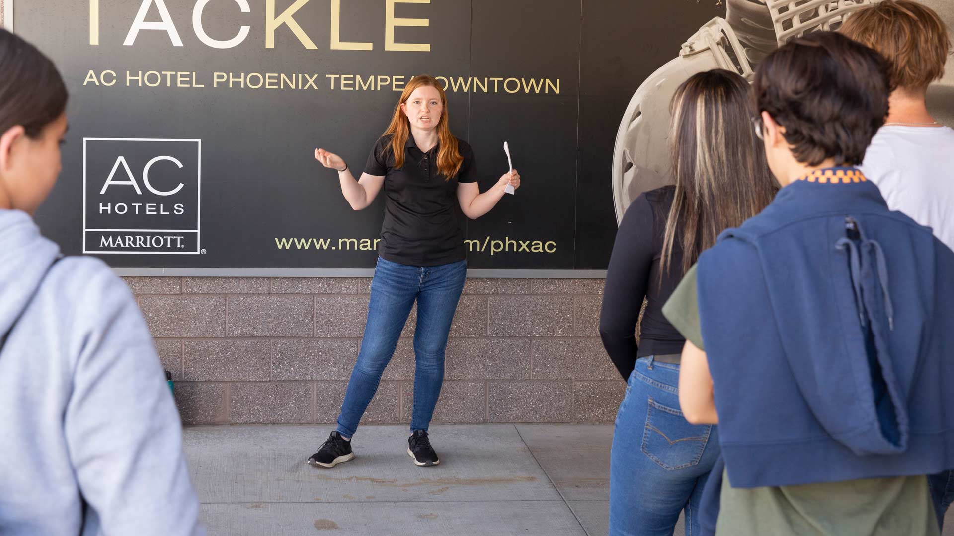 Students listen to female volunteer talk about engineering under concrete overhang at Sun Devil Stadium