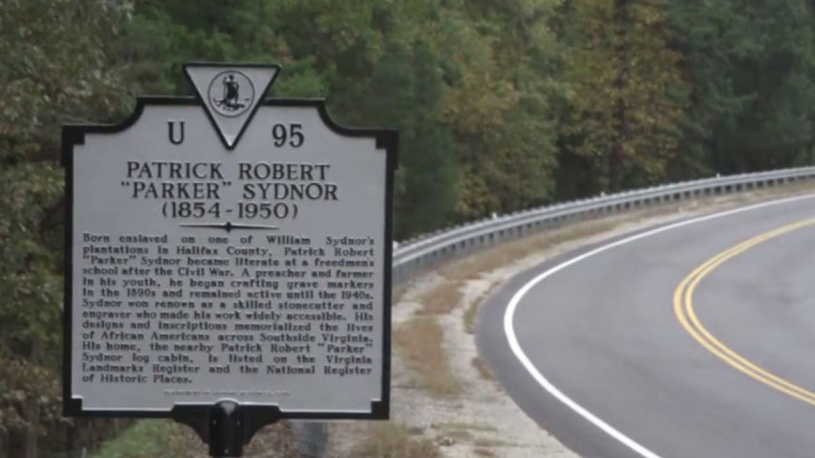 A historical-site highway marker stands next to a wooded road.