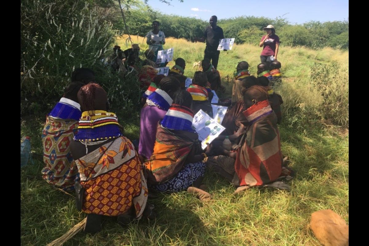 Field researchers speaking to people seated in a grassy area, wearing brightly colored fabrics.