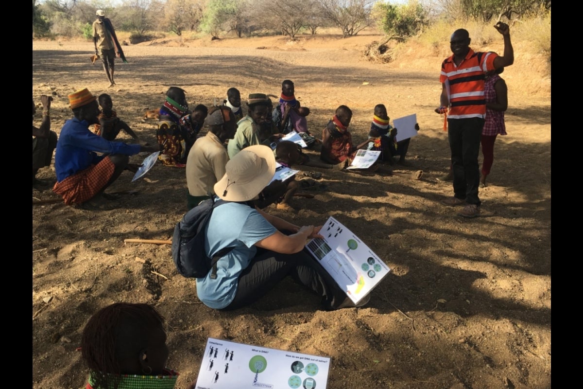 People seated in a sandy outdoor area listening to a standing man speak.