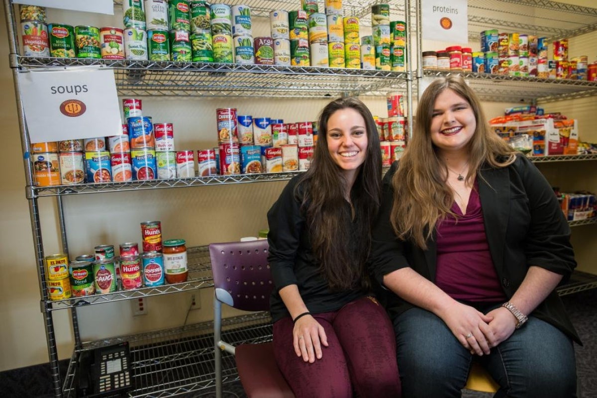 two women seated in front of shelves of canned food