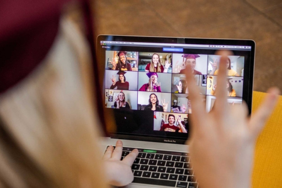 A woman wearing a graduation cap looks at a laptop Zoom screen full of other graduates