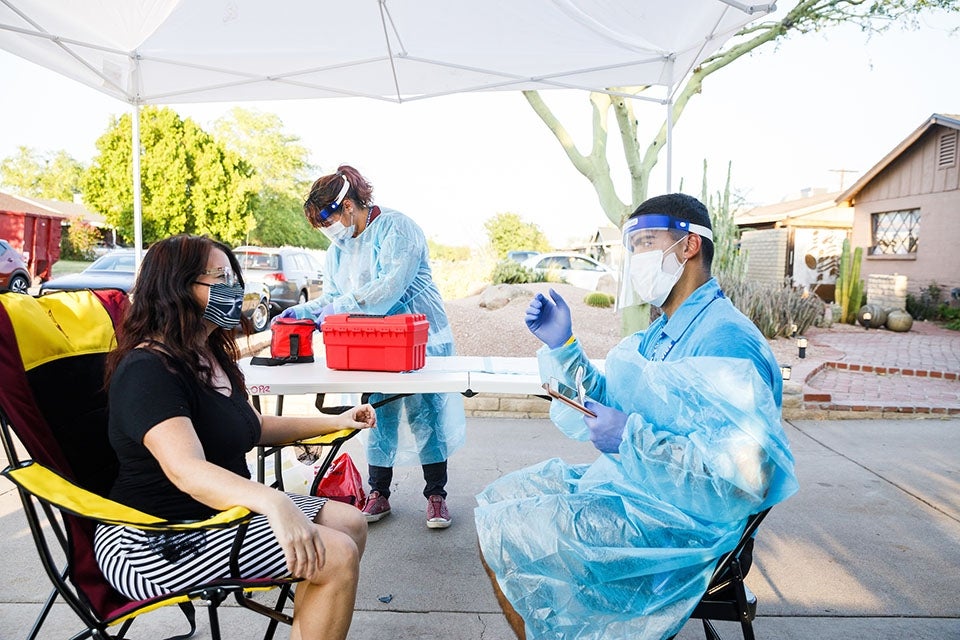 A man in full personal protective gear, tablet in hand, speaks to a woman beneath a collapsible tent in a driveway.