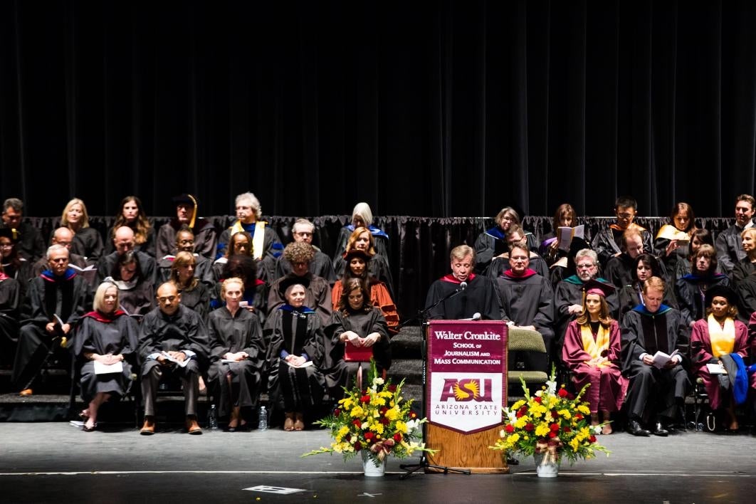 man speaking on stage with graduates behind him