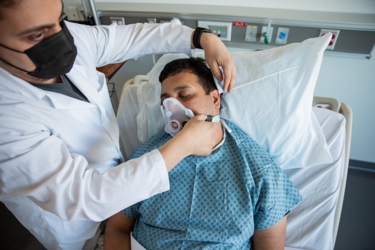 A man in a lab coat hooks a breathing device onto a man on a demonstration hospital bed