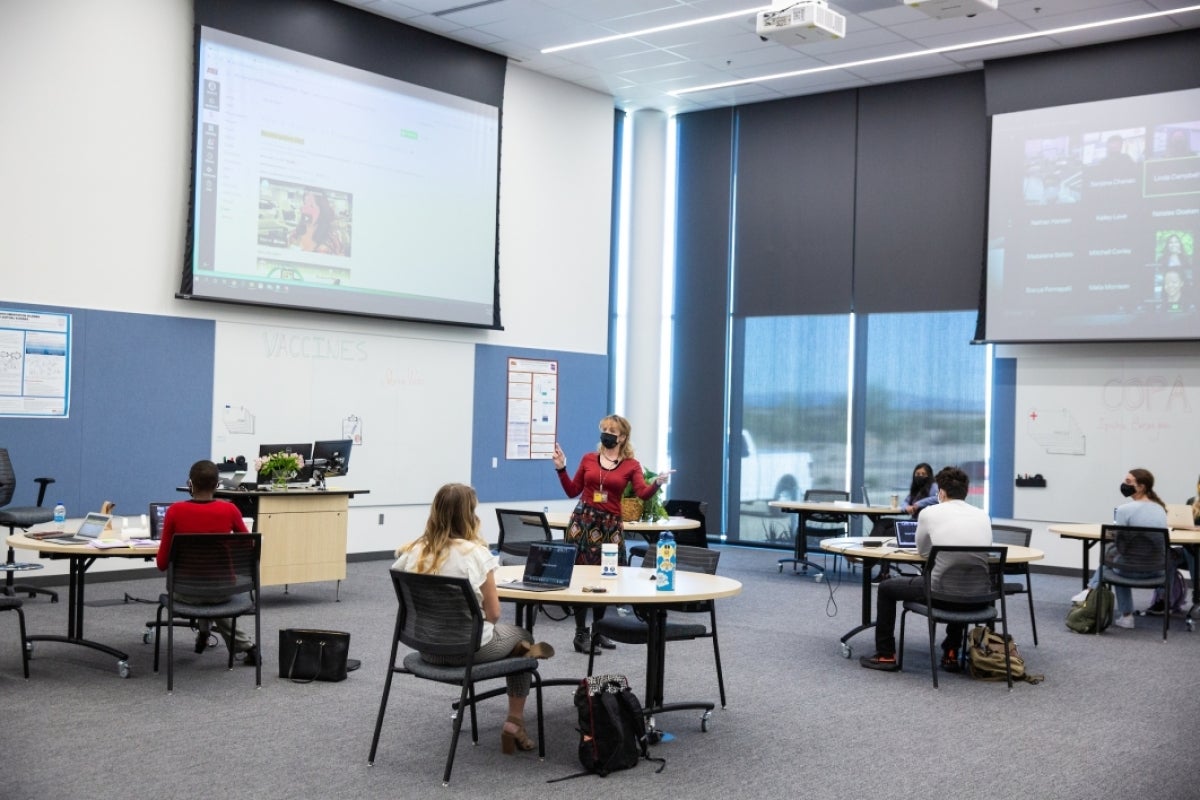 Students sit distanced at tables while a professor leads a class, with large screens showing others on Zoom