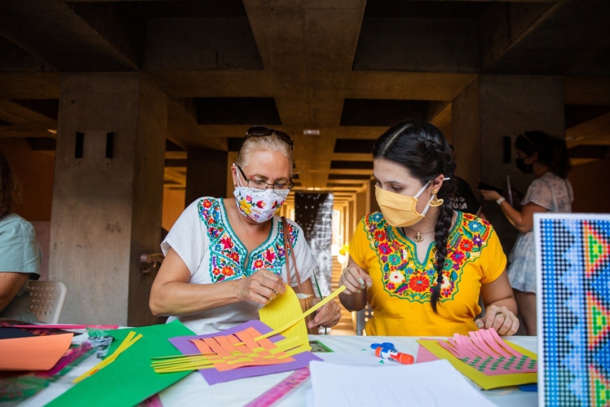 Two women wearing blouses with Mexican embroider sit at a table and work on paper weavings