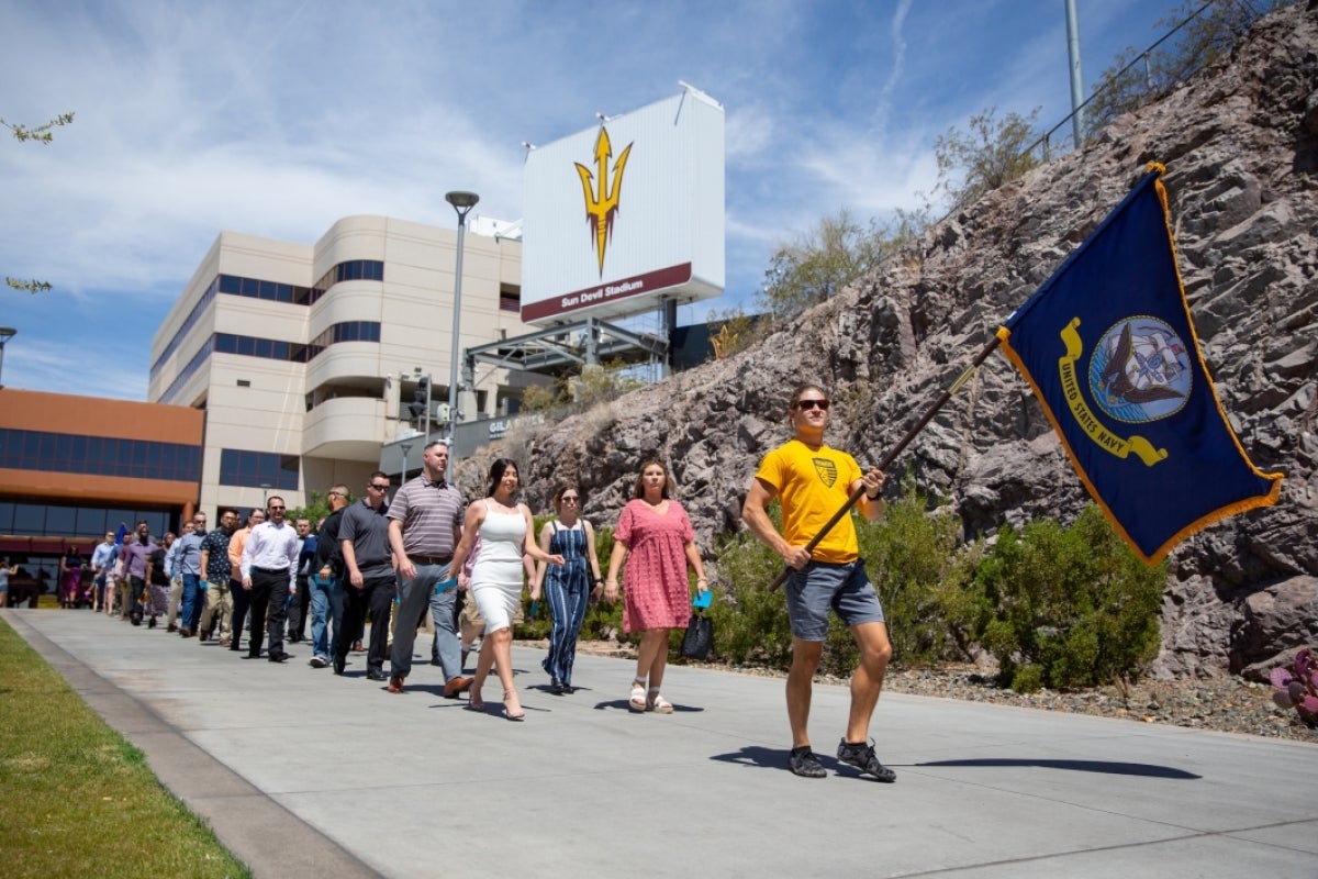 A man holds a Navy flag while veterans walk in a line behind him