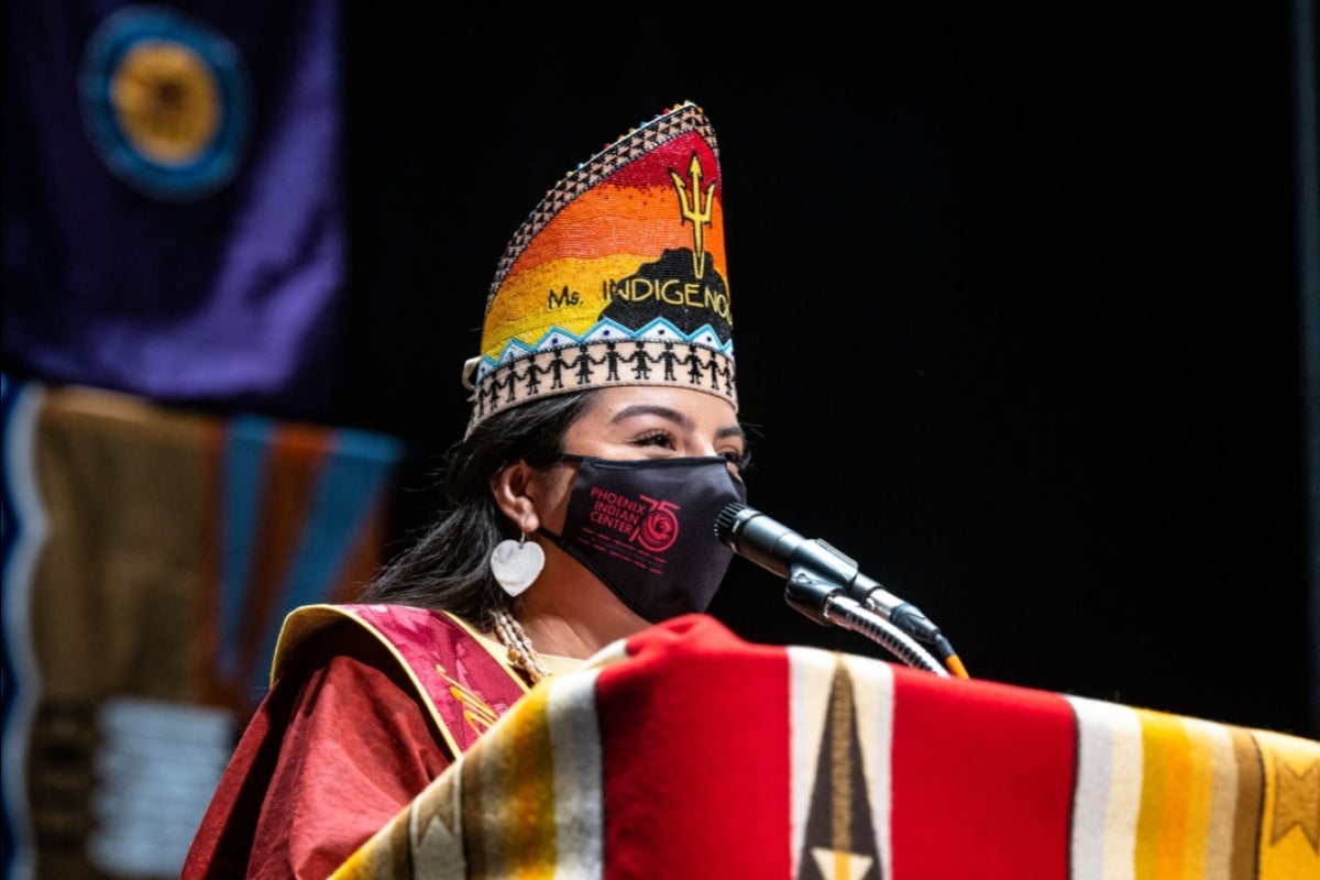 A woman in a Native American crown speaks at a lectern while wearing a mask