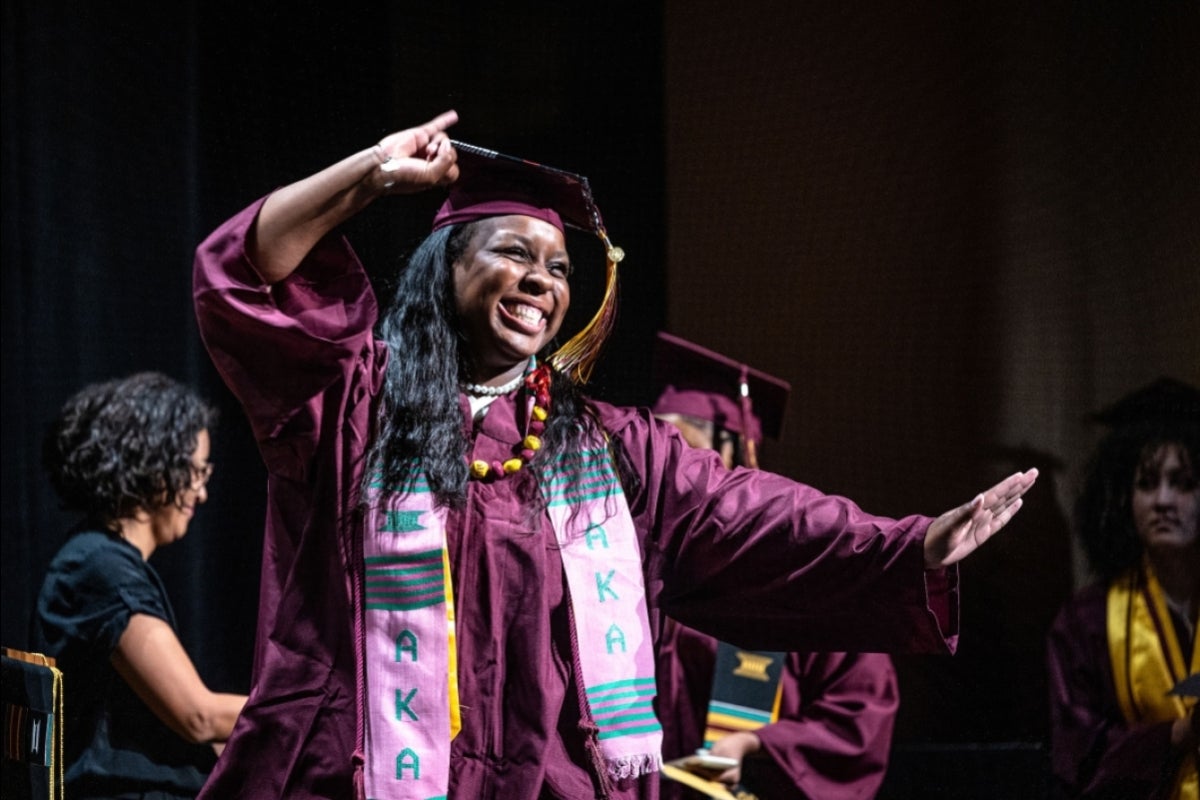 A woman in graduation regalia dances across a stage to receive a stole