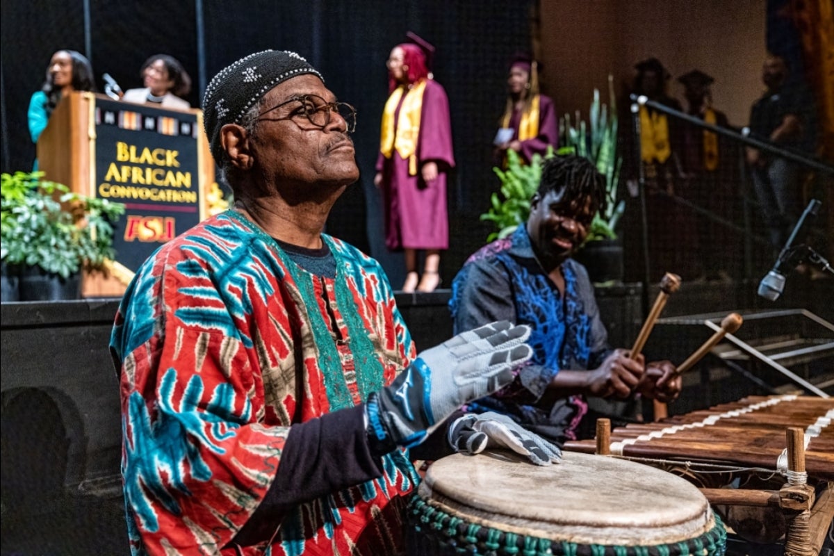 A man plays a large bongo-type drum in the foreground while another man playes a xylophone in the background