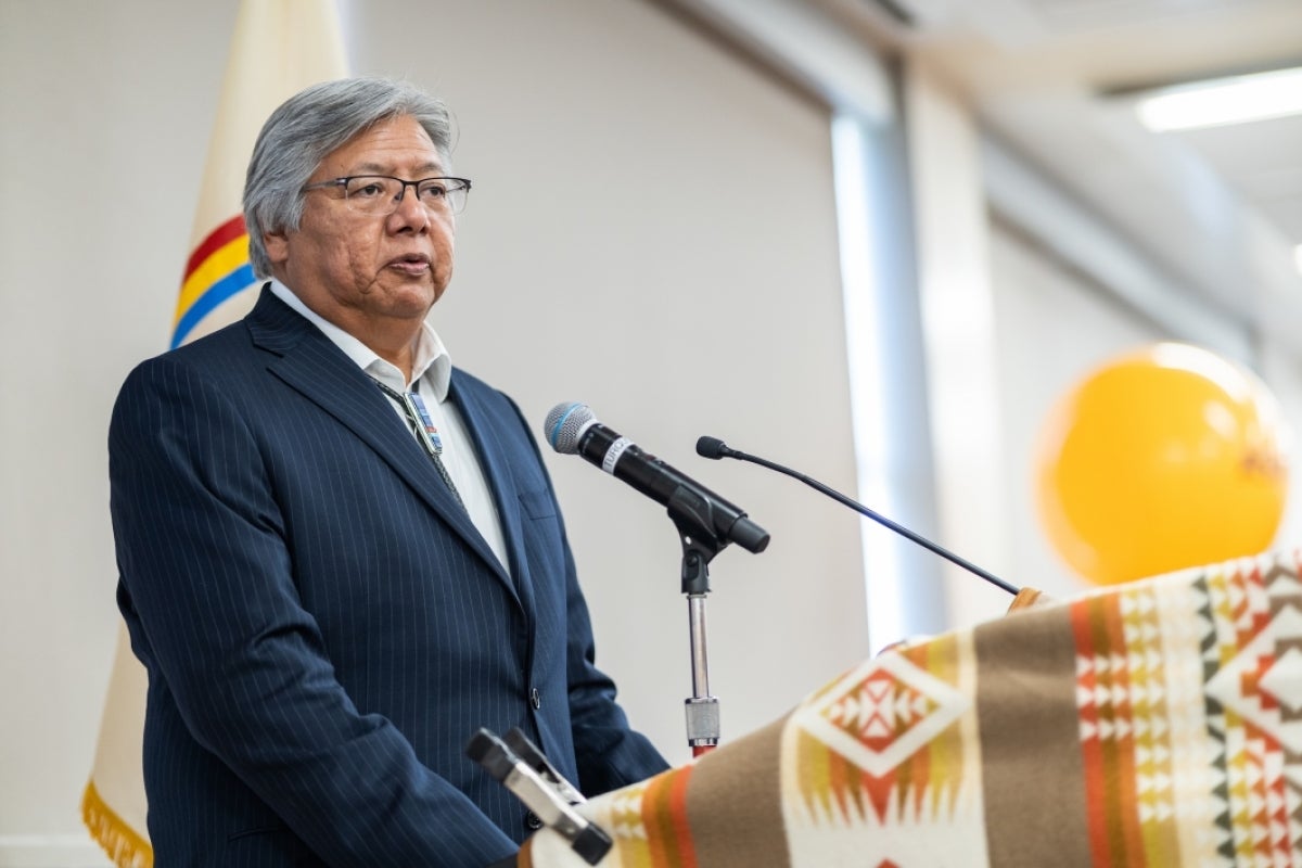 A man speaks at a lectern covered in a Native American textile with a memorial photo on an easel to his right