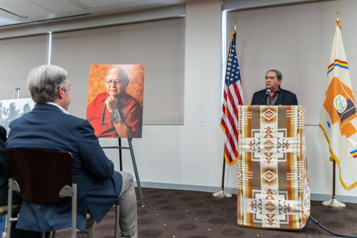 A man speaks at a lectern covered in a Native American textile with a memorial photo on an easel to his right