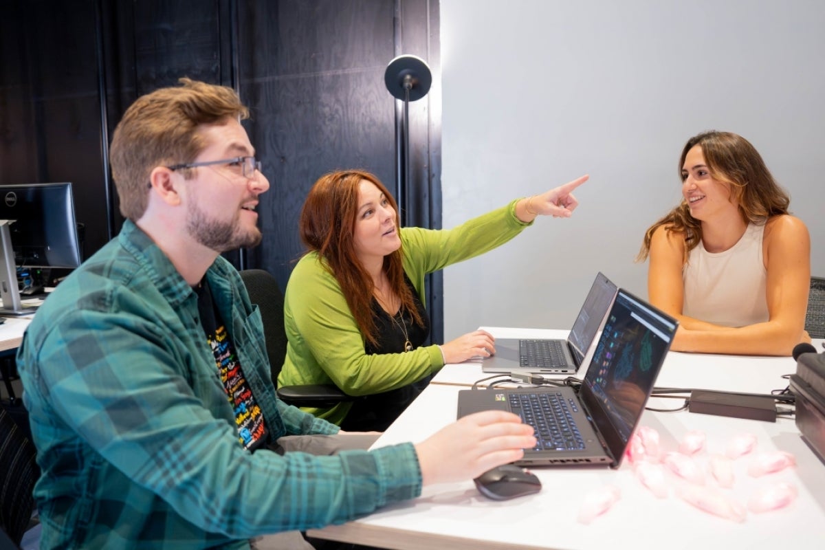 Three people sit at a table while one of them points at a screen off camera