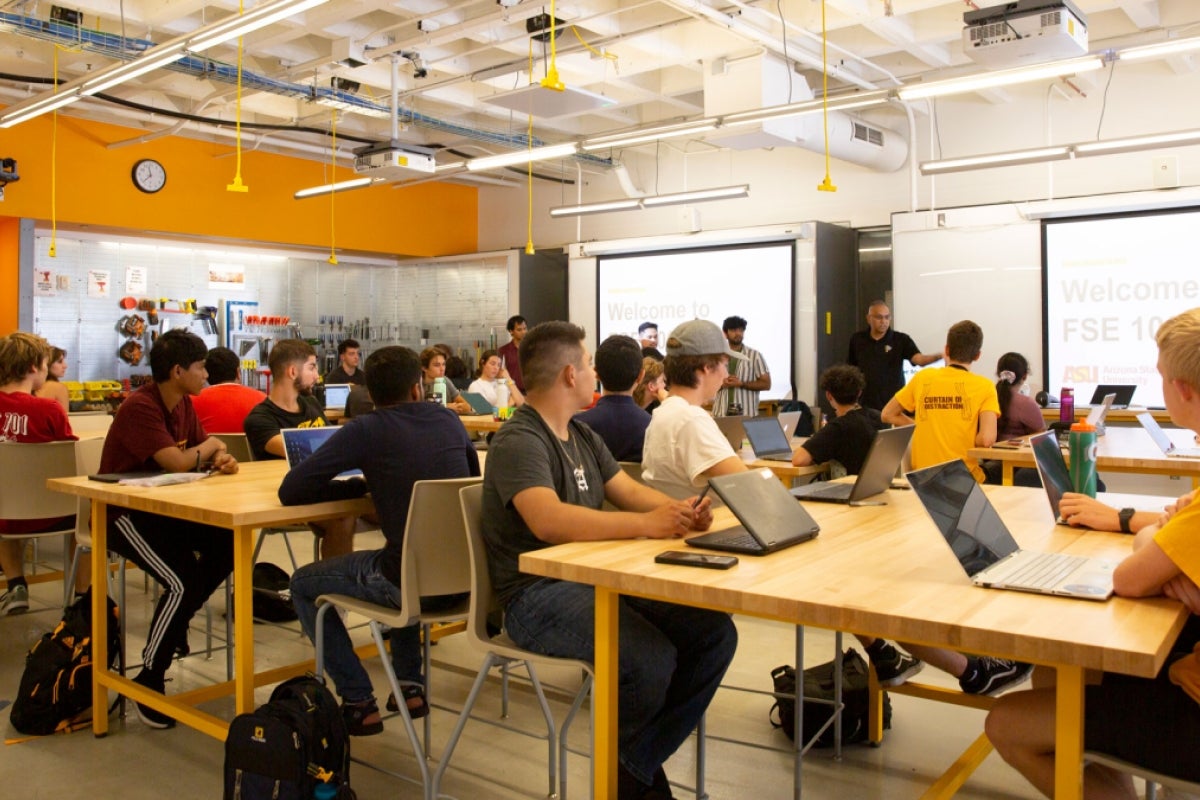 A classroom full of students sitting at tables with laptops watch as a professor presents to the class.