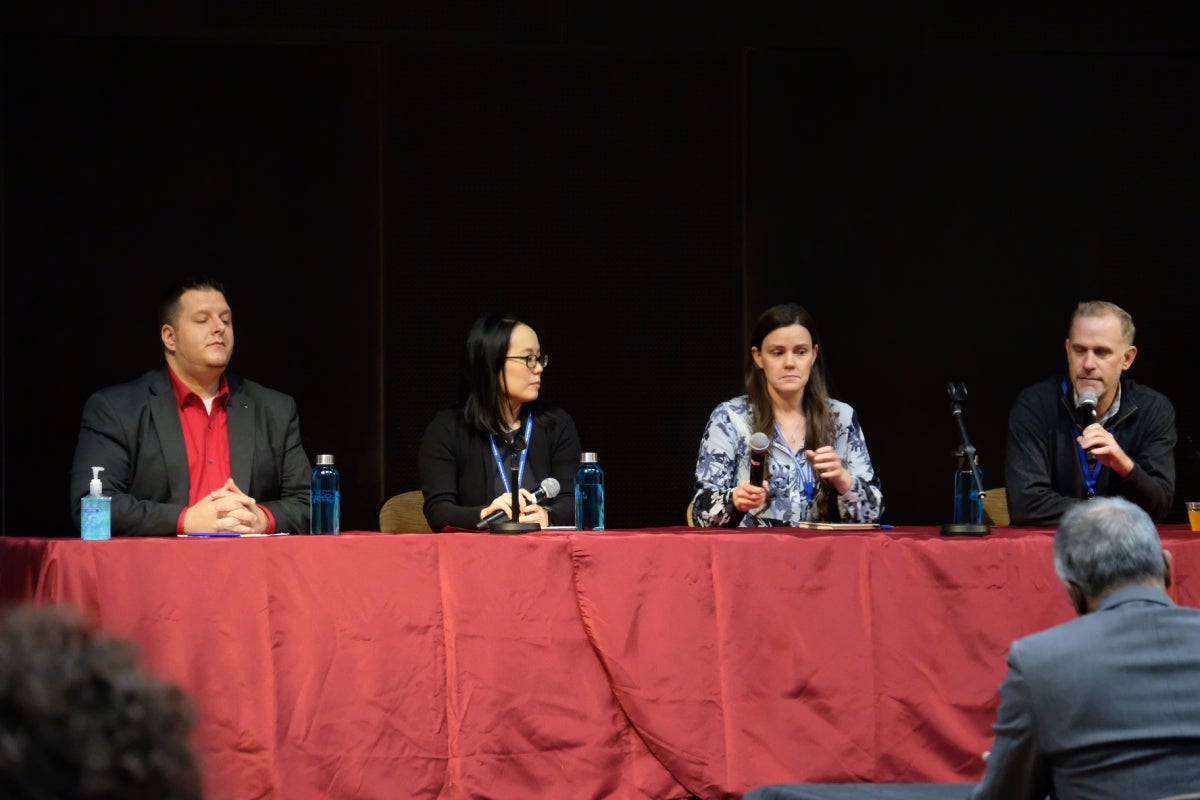 Four panelists sit in a line at a table in front of a crowd