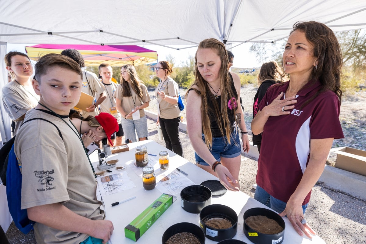 Two women stand behind a table where kids are observing items displayed