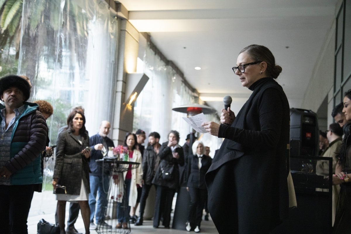 Woman speaking into a microphone as people in a museum lobby look on.