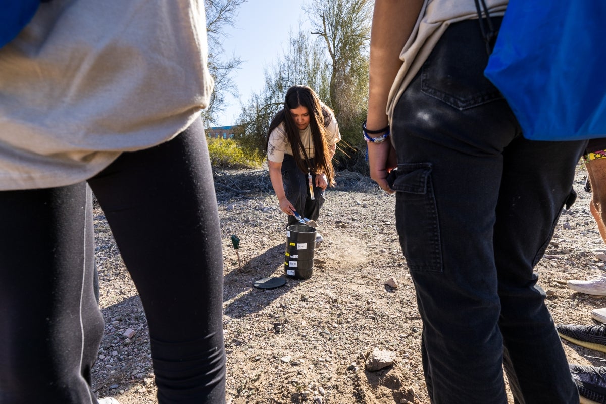 A woman stands in front of kids using a dirt sifter