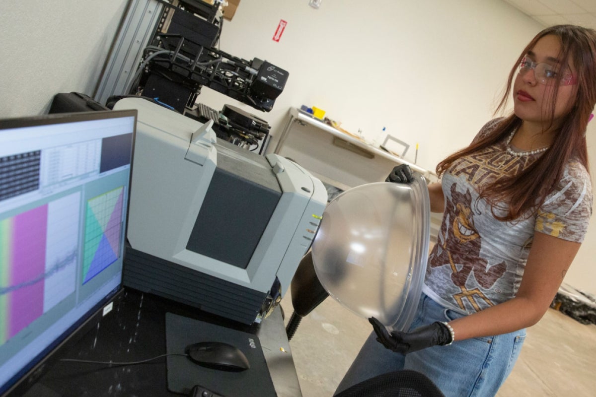 A woman holds a spacesuit helmet bubble while looking at machinery