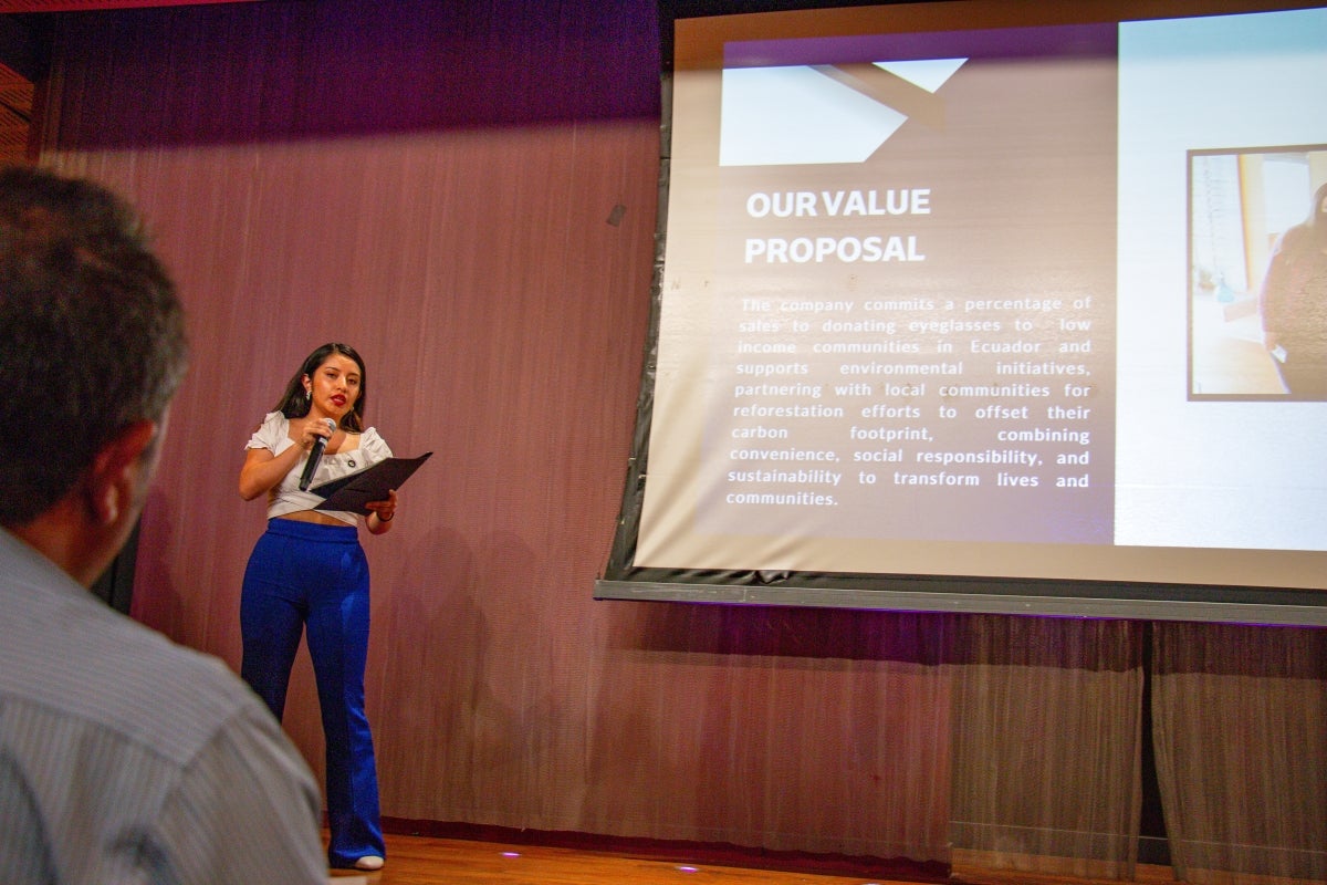 Woman speaking into a microphone in front of a screen with a presentation slide projected onto it.