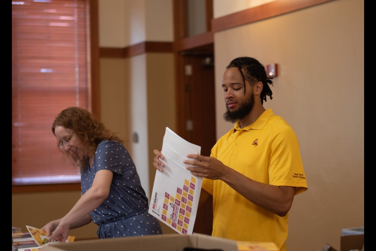 Two people assembling resources in an envelope, one is a woman in a blue polka dot dress, and the other is a man in a yellow polo shirt.