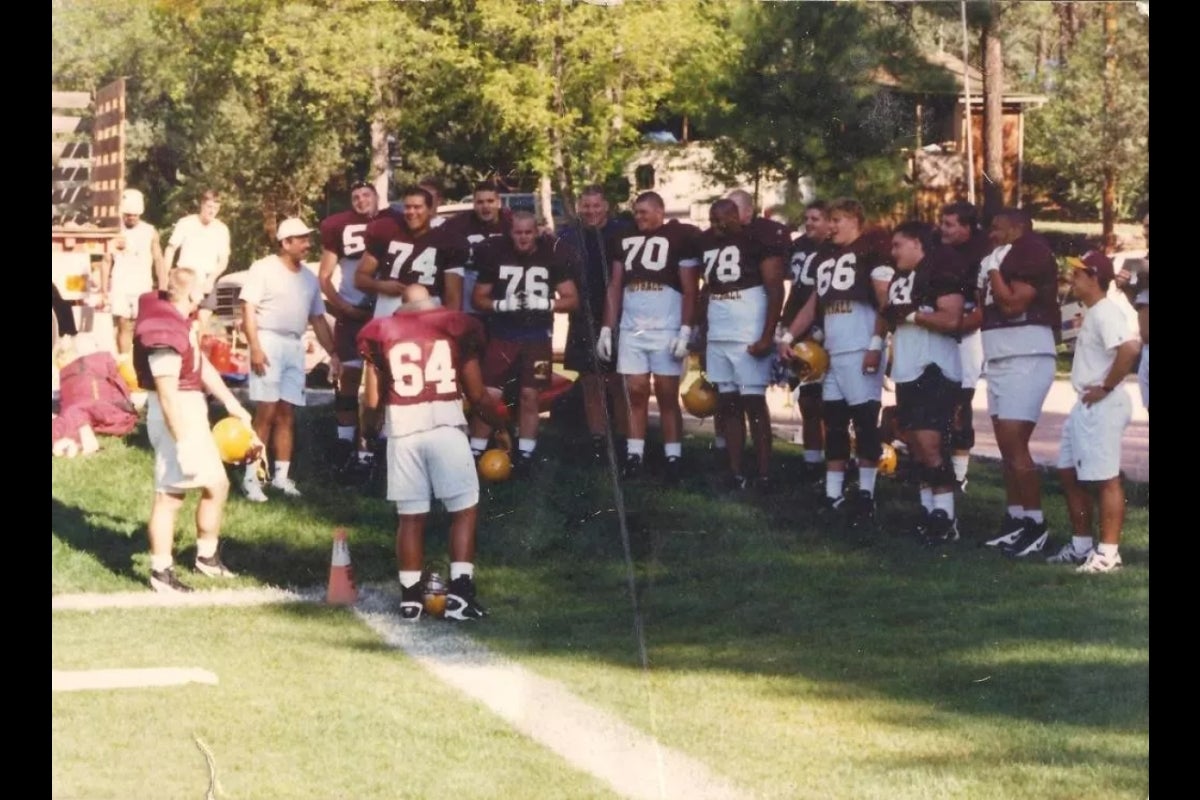 ASU football team from the 1990s hanging out on a practice field surrounded by pine trees