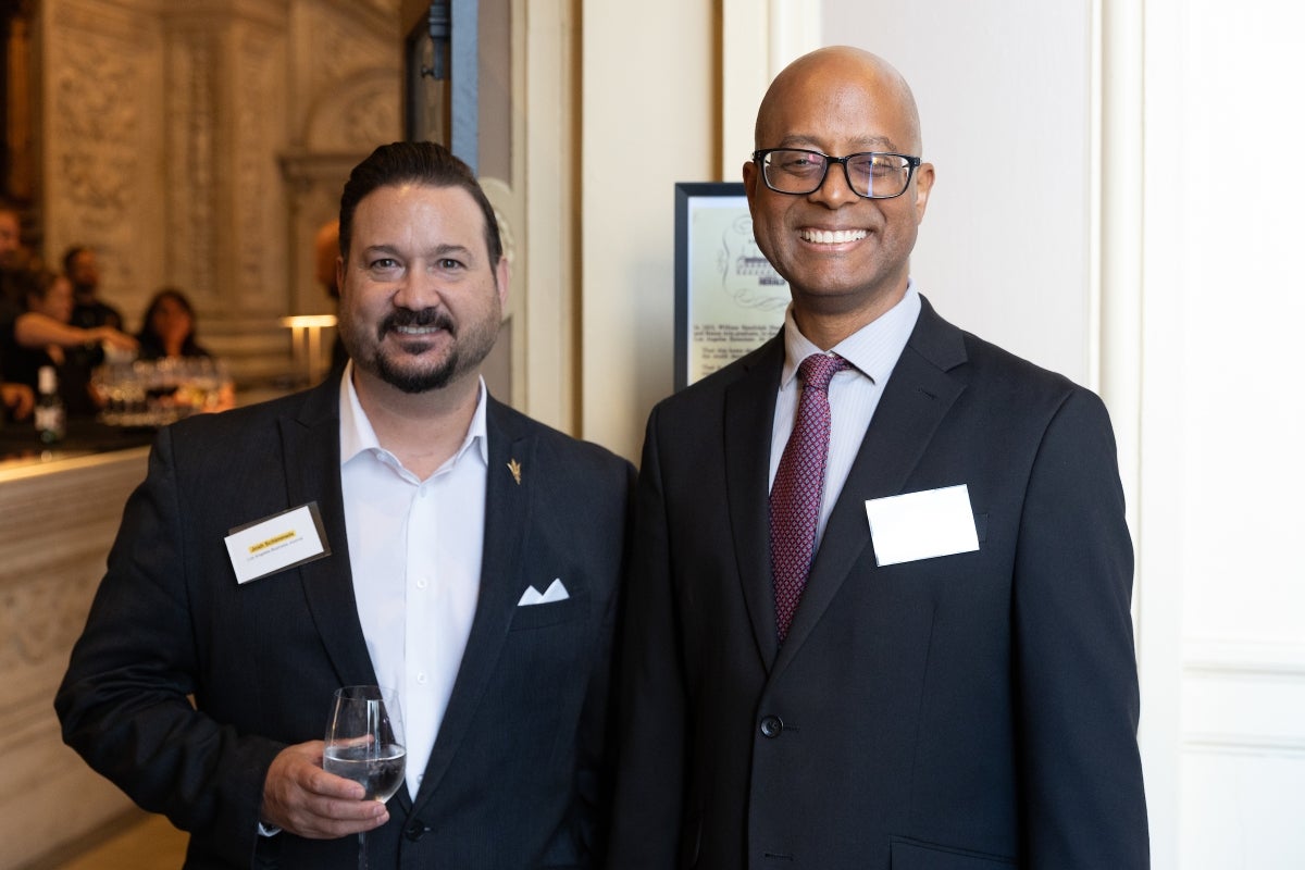 Two men in dark suit jackets pose for photo