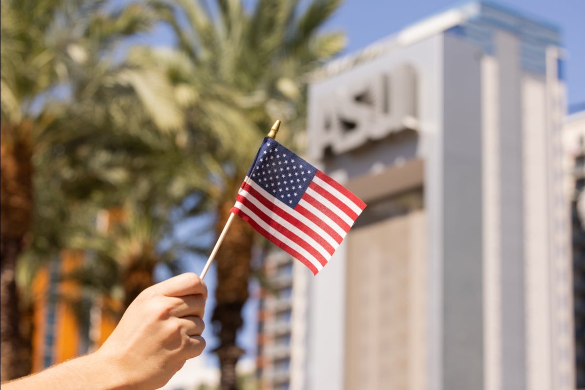 A hand holds a tiny American flag in front of an ASU sign