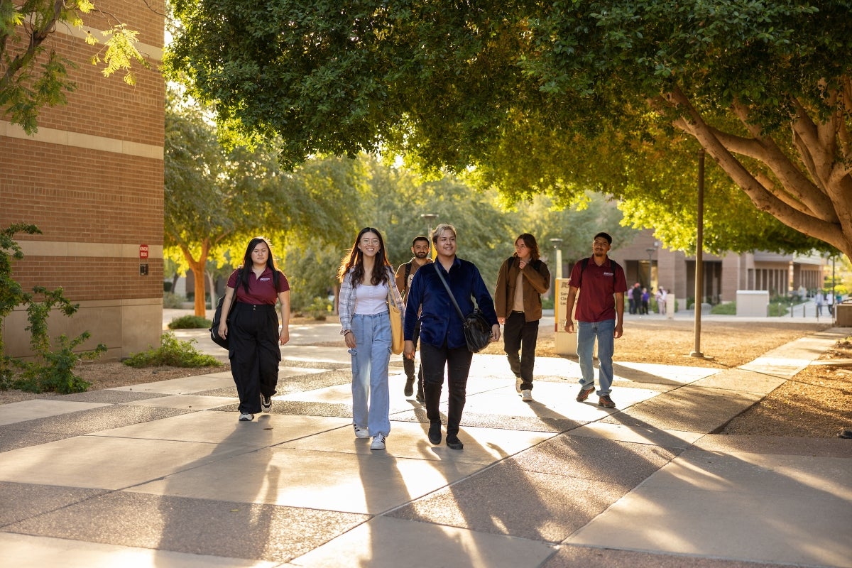 A group of students walk on a tree-lined walkway on ASU's west campus with the sun setting behind them