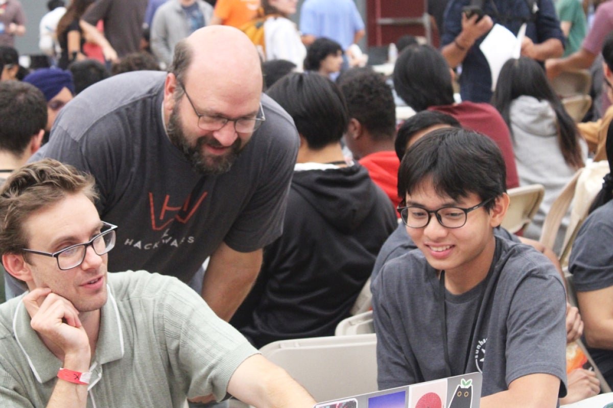 Three students sit around a laptop in a packed room.