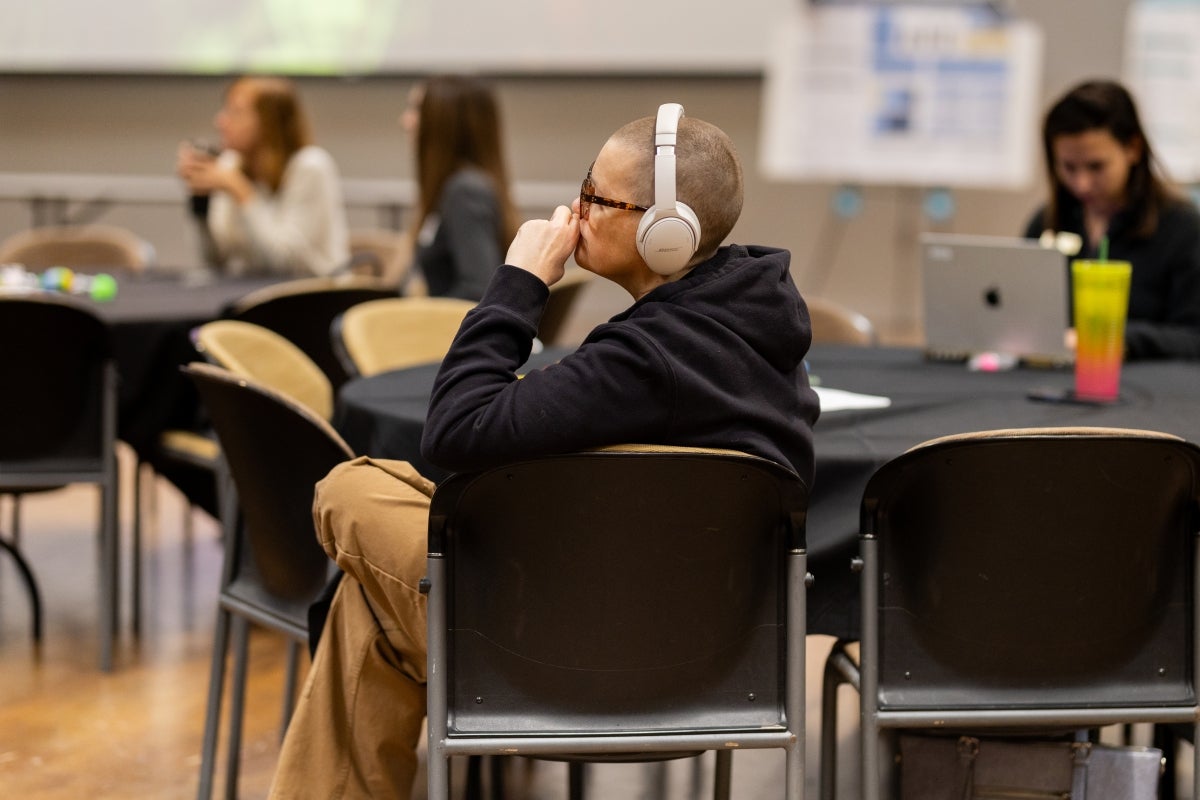 Person in black jacket is sitting at table with white headphones on looking ahead