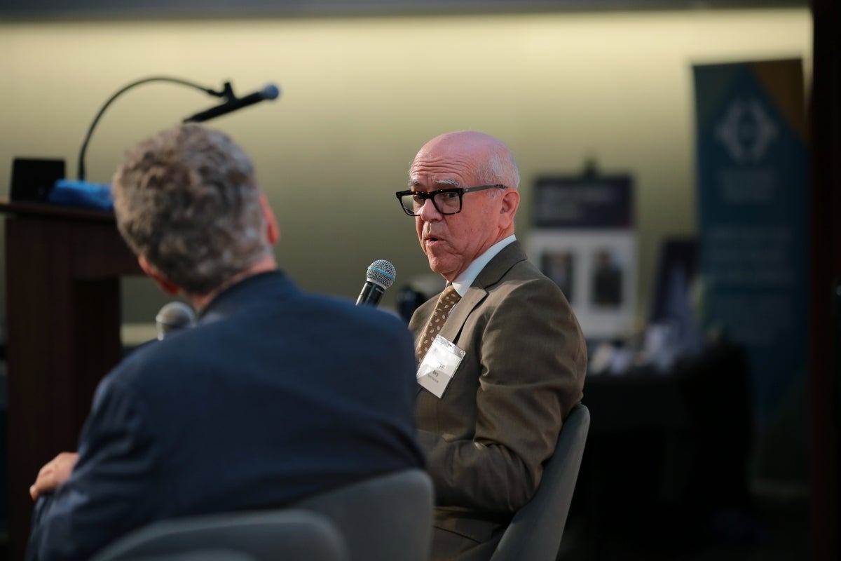 Older man with black glasses wearing a brown suit jacket speaks to another person on stage for a panel