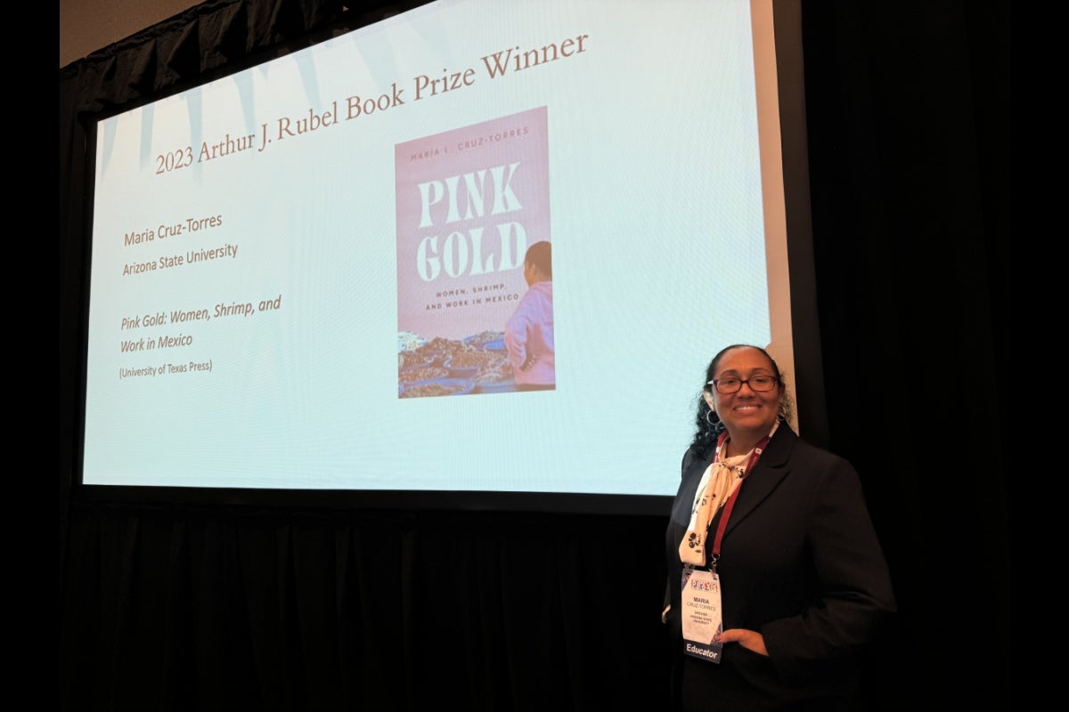 ASU Associate Professor Maria Cruz-Torres stands next to a projector screen announcing her book award