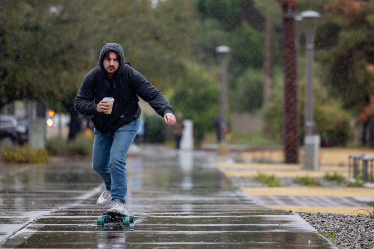 A man rides a skateboard in the rain while holding a cup of coffee