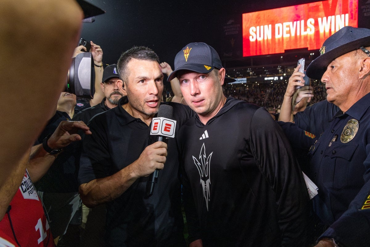 A reporter holding a mic with an ESPN label interviews a coach on a football field with a screen behind that reads: Sun Devils Win