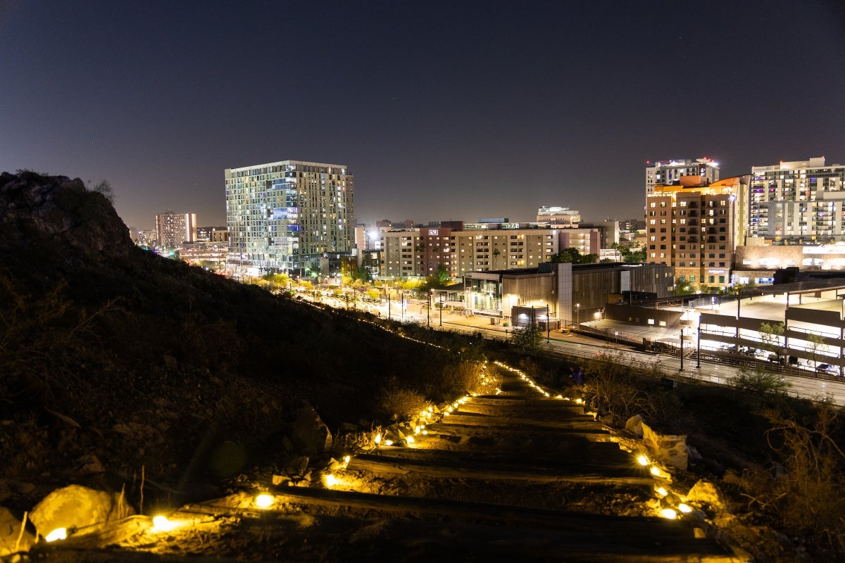 A trail on a mountain is lined with lights, as the nighttime skyline of the city rises behind it