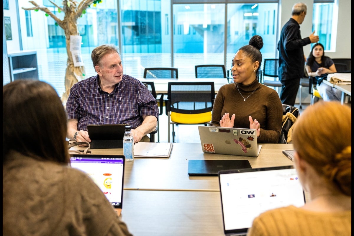 Group of students of all ages seated at a table talking and working on laptops