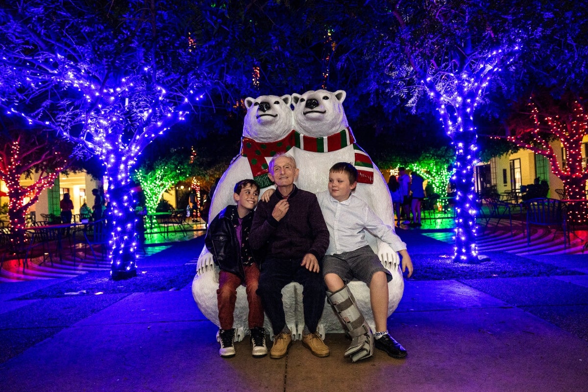 A grandpa and two grandsons sit on a bench made out of two polar bears with trees behind them full of Christmas lights