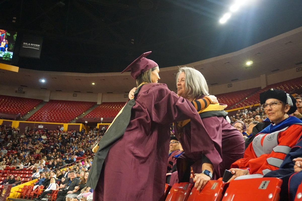 Student and professor wearing graduation regalia hug in a large arena.