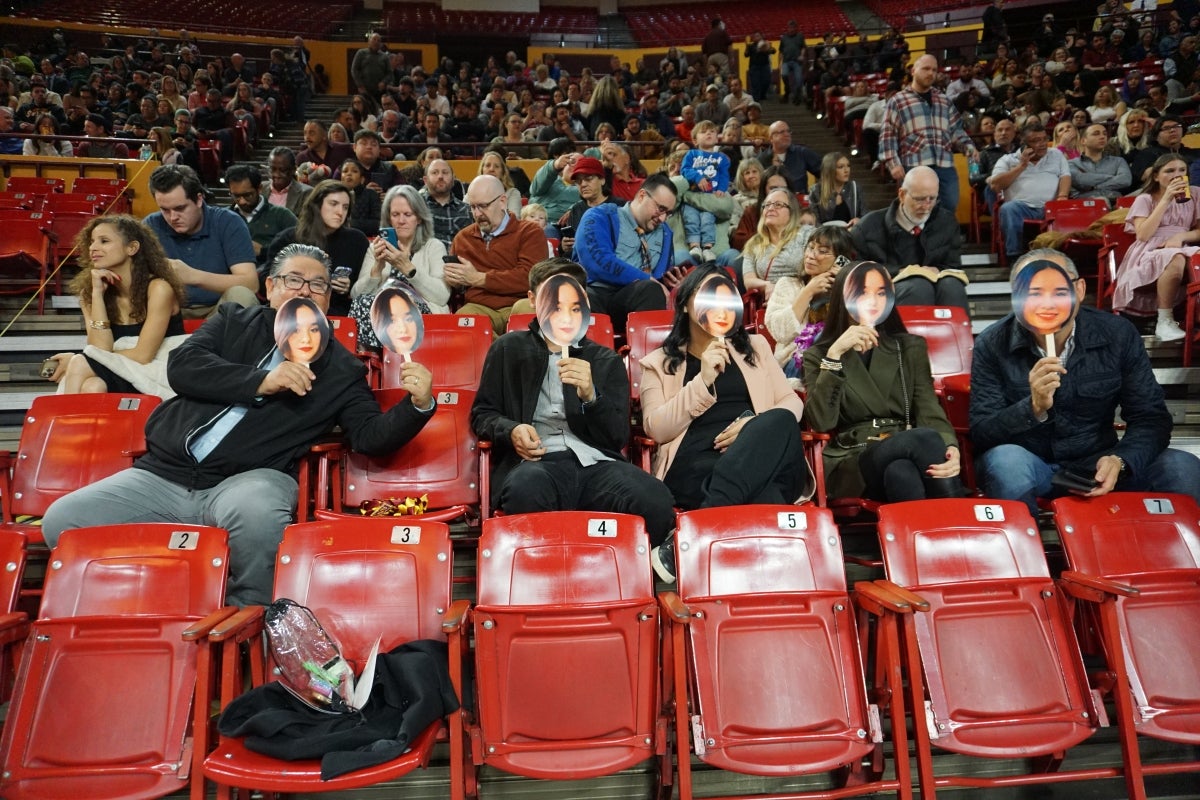 People seated in an arena holding up photos of a graduate in front of their faces.