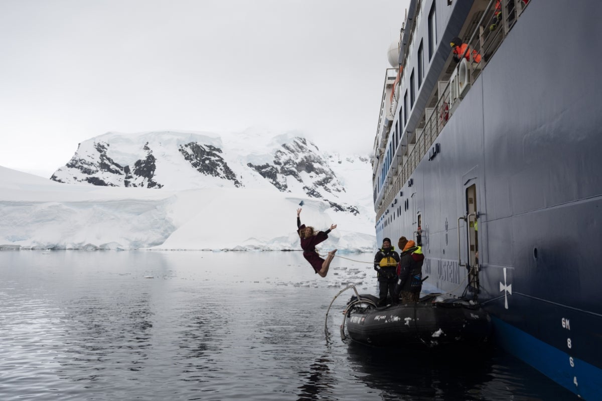 Woman jumping from a large boat into the ocean with a snowy, rocky landscape in the background.