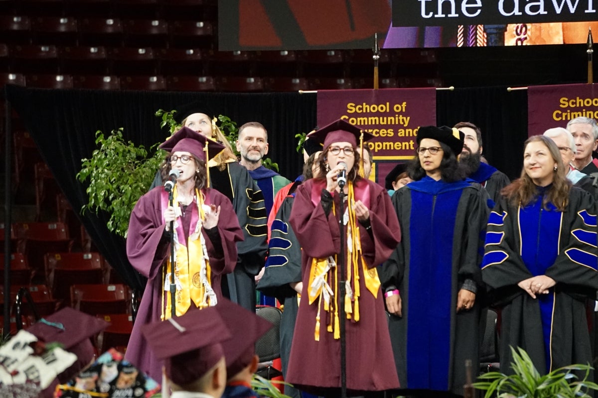 Students wearing graduation regalia singing onstage at a ceremony.