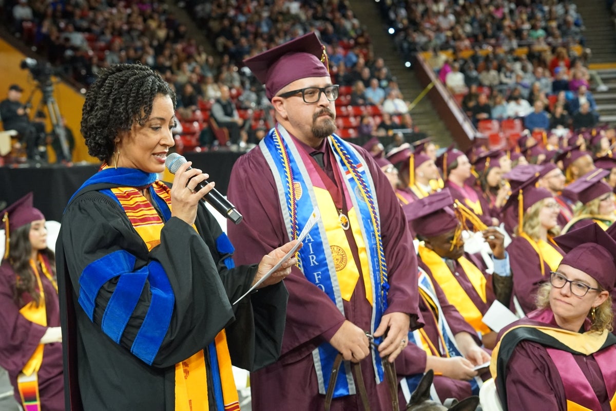 Woman wearing graduation regalia addresses students at a graduation ceremony.