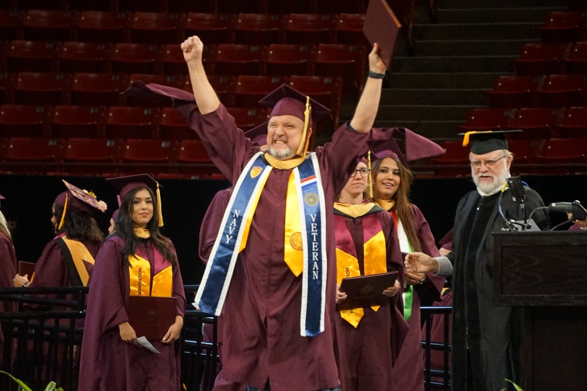 Man in maroon cap and gown raising both hands onstage.