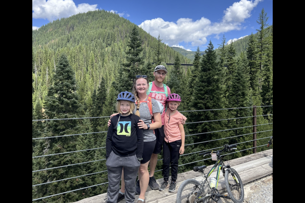 Two adults and two children wearing biking gear pose next to a railing in front of a scenic tree-lined view.