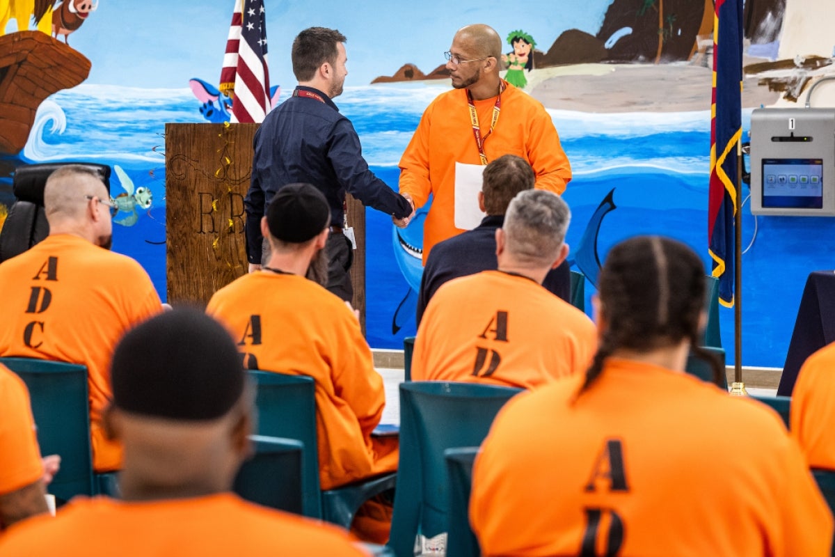 Man in blue button down shirt shakes hands with inmate in front of crowd of other inmates wearing orange jumpsuits