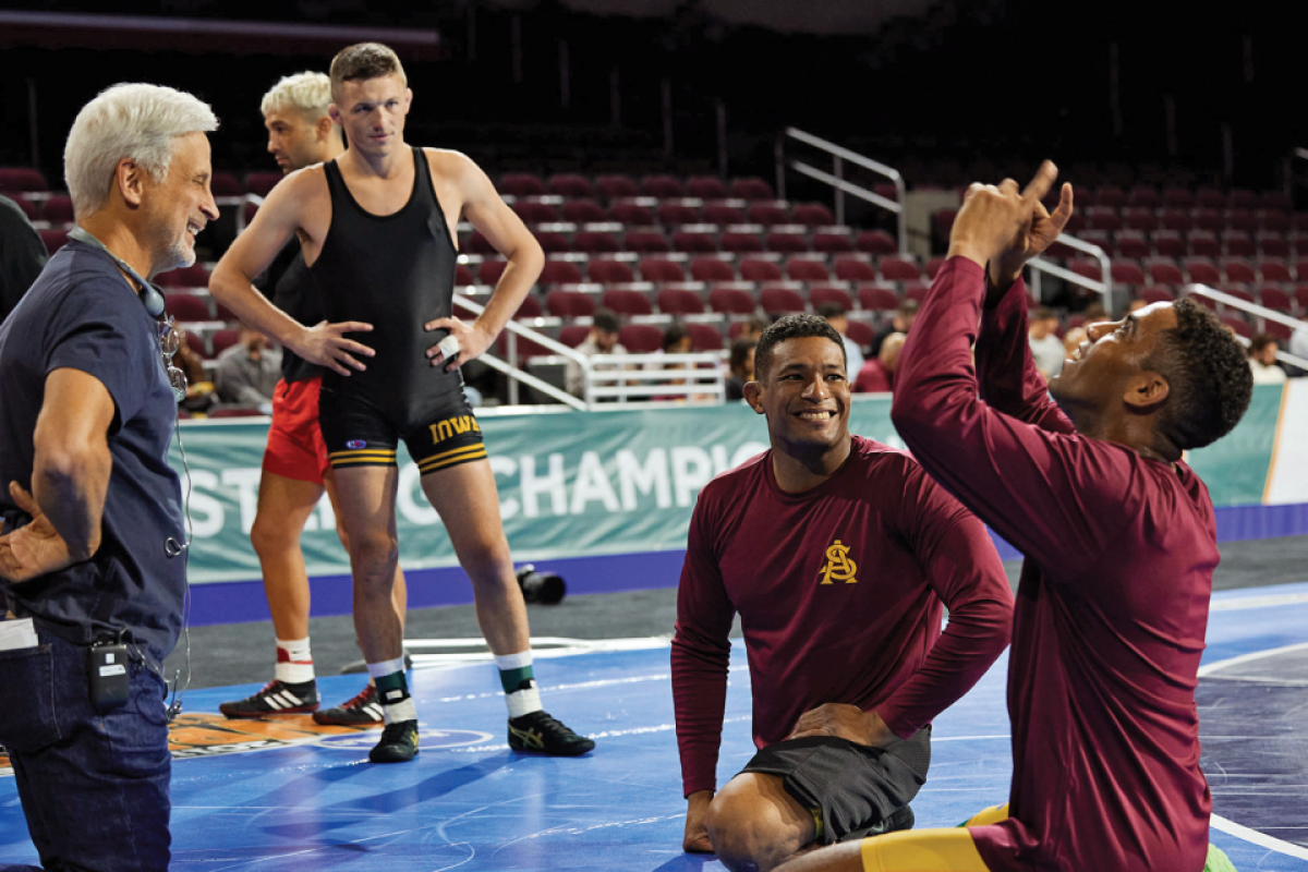 People stand and sit on a wrestling mat during a film shoot