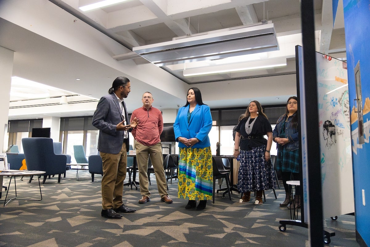 A group of five people engaged in a discussion in a modern library setting with patterned carpet and lounge furniture.