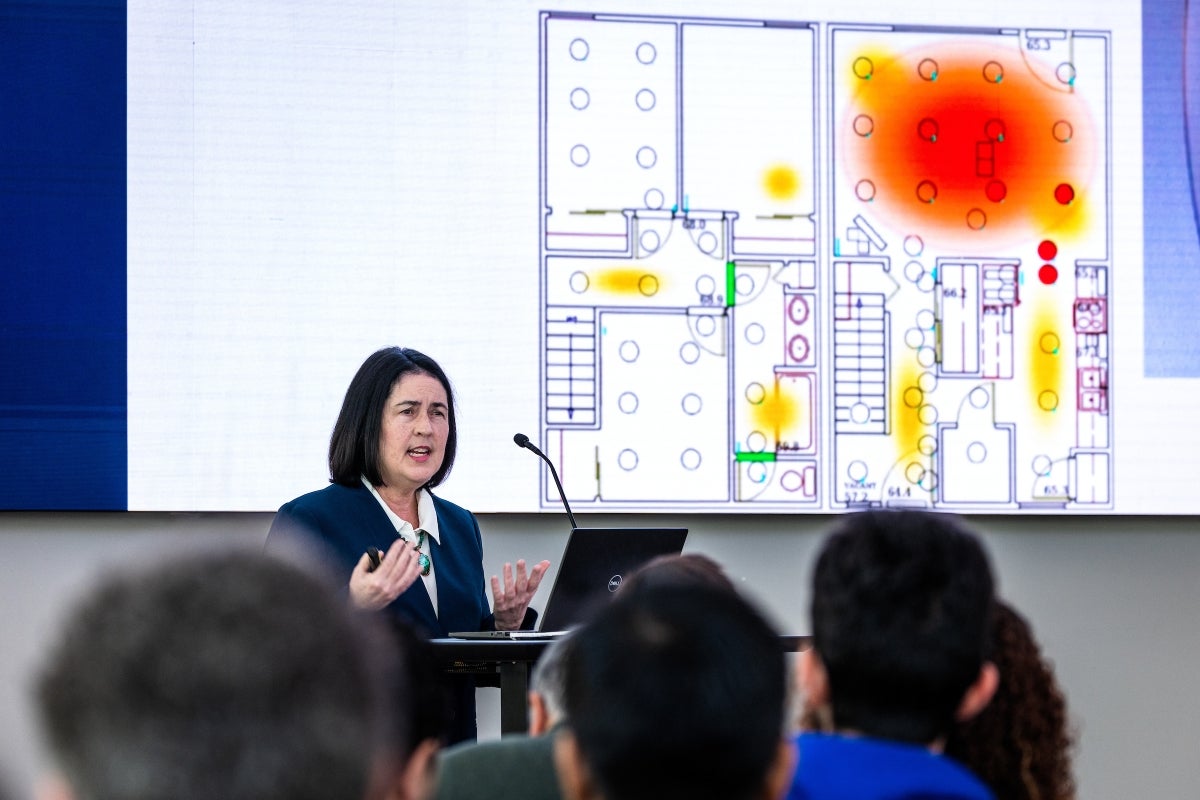 A woman talks into a microphone in front of a screen displaying a blueprint of a building.